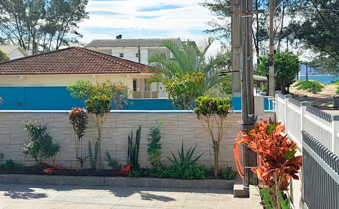 Front yard with a low stone wall, landscaped garden bed, and tall trees; blue fence and a path toward the beach visible in the distance.