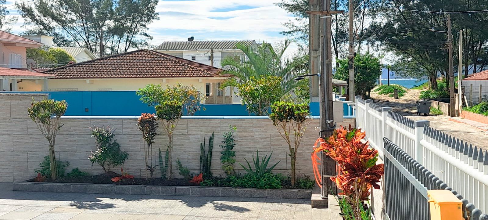 ritaf2 Front yard with a low stone wall, landscaped garden bed, and tall trees; blue fence and a path toward the beach visible in the distance.
