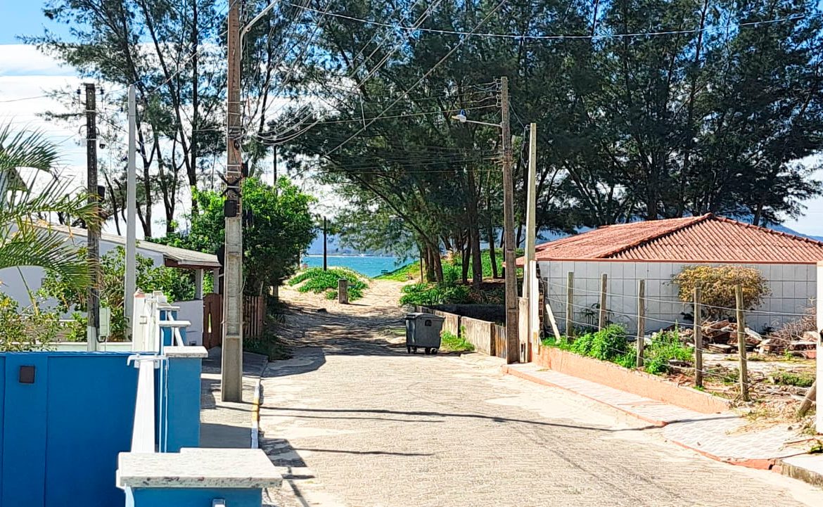 Street in a sunny coastal neighborhood leading to the beach, with a blue wall on the left and trees lining the road.